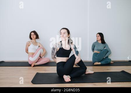 Ruhige, leicht gelangweilte Frauen in Sportbekleidung üben Yoga in einer Gruppe, machen seitliche Verdrehungen in einem großen Studio. Konzentriere dich auf ein Mädchen mit großer Schulter Stockfoto