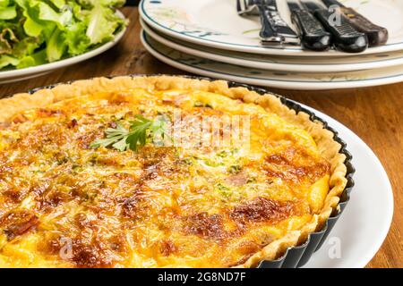 Hochwinkelansicht von hausgemachtem Speck Spinat Quiche in schwarzem Metall Ofen Kuchen Backplatte auf weißer Keramikschale mit grünem Gemüse, Stapel von weißem Ceran Stockfoto