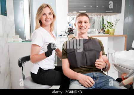 Fröhliche junge Kieferorthopäde, die die Kamera anschaut und lächelt, während ein männlicher Patient mit Zahnspangen die Daumen hoch gibt. Konzept der Zahnmedizin, zahnärztliche Stockfoto
