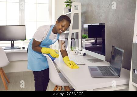 Fröhlicher Mitarbeiter vom Hausmeisterdienst, der Bürotische und Tische mit Wischtuch putzte Stockfoto