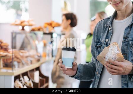 Bokeh-Bild eines Mannes in einer Jeansjacke, der eine Tasse Kaffee und ein Croissant in einer Papierverpackung hält. Abgeschnitten, Halbgesicht, lächelnd. Stockfoto