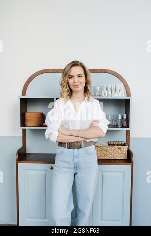 Selbstbewusste blonde Frau in den Dreißigern, die mit gekreuzten Händen vor dem Küchenschrank steht. Sie trägt ein Hemd und eine leichte Jeans und schaut sich an Stockfoto