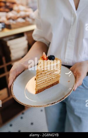 Ein Stück eines russischen Honigkuchens auf einem Teller, den die Frau hält. Mit Beeren verziert, zerig hoch und geschichtet. Stockfoto