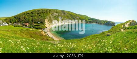 Das klare Wasser der Bucht Lulworth mit dem südwestlichen Küstenpfad auf dem Hügel an einem hellen Sommertag mit Gras und Blumen, die zum Strand führen. West Stockfoto