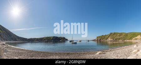 Weit entfernt von der Bucht Lulworth, wo die Morgensonne die Kieselsteine und Felsen am Strand erleuchtet, mit Segelbooten, die in der Bucht, West Lulworth, verankert sind, Stockfoto