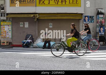 Eine obdachlose Person sitzt auf ihrer Sammlung von recyceltem Material, während zwei Frauen in einer Seitenstraße in Shinjuku, Tokio, Japan, Fahrrad fahren. Stockfoto