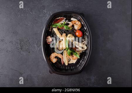 Schwarze Spaghetti Nero mit Meeresfrüchten, Tomaten und Basilikum in schwarzem Kunststoffbehälter auf dunklem Hintergrund. Designer-Food-Konzept für die Mitnahme oder Lieferung Stockfoto