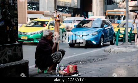 Der traditionelle thailändische Old man Street Musiker Siam Square Wirtschaft und Wirtschaft Bangkok Thailand spielt auf einem Hocker um Veränderung Stockfoto