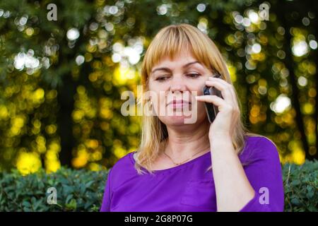 Unschärfe kaukasisch blonde Frau reden, sprechen am Telefon draußen, im Freien. 40-jährige Frau in lila Bluse im Park. Erwachsene Frauen, die ein Mobiltelefon verwenden Stockfoto
