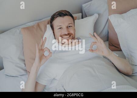 Entspannter junger bärtiger Mann, der in seinem Bett lag und ein Schild mit OK zeigte Stockfoto