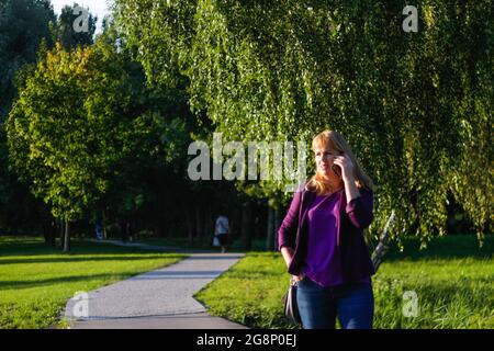 Unschärfe kaukasisch blonde Frau sprechen auf dem Telefon draußen, draußen. 40-jährige Frau in lila Bluse im Park stehend. Erwachsene Frauen, die Telephon benutzen Stockfoto