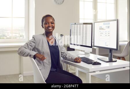 Glückliche schwarze Geschäftsfrau oder Buchhalterin, die am Schreibtisch mit Desktop-Computern sitzt Stockfoto