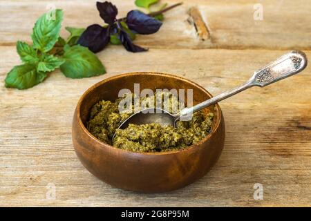 Basilikum-Pesto in Bambusschüssel mit Metalllöffel und grün-violetten Blättern auf Holztischhintergrund Stockfoto