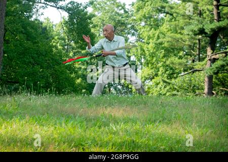 Ein älterer asiatischer Amerikaner, wahrscheinlich Chinese, macht Tai-Chi-Übungen mit einem Schwert. Auf einem Hügel in einem Park in Queens, New York City. Stockfoto