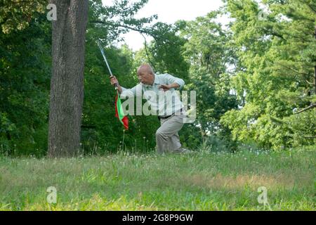 Ein älterer asiatischer Amerikaner, wahrscheinlich Chinese, macht Tai-Chi-Übungen mit einem Schwert. Auf einem Hügel in einem Park in Queens, New York City. Stockfoto