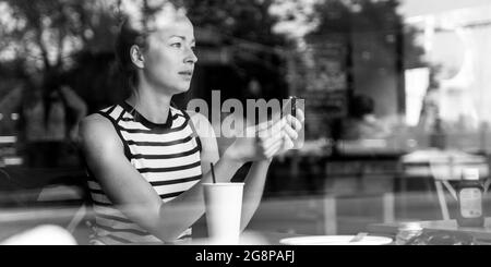 Nachdenklich kaukasische Frau mit Mobiltelefon, während Sie durch den Coffee shop Fenster während der Kaffeepause. Stockfoto