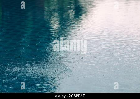 Regen fällt auf die Wasseroberfläche in einer Pfütze mit abgestuftem Schatten von schwarzem Schatten und Reflexion des blauen Himmels Stockfoto
