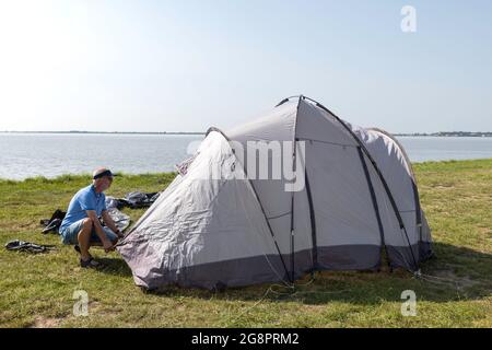 Reifer Mann, der ein Zelt aufstellt und auf einem Feld nahe dem Wasser auf einem schönen Platz nahe dem See Zeltpfeifer ablegt Stockfoto