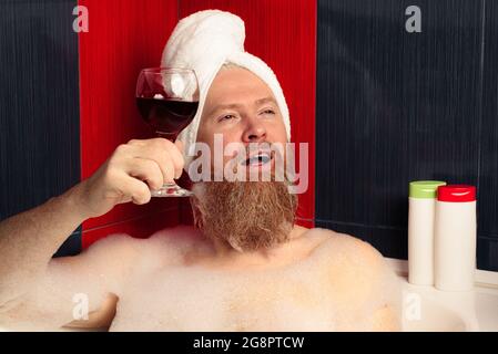 Glücklicher bärtiger Mann, der ein Schaumbad nahm. Lustiger Typ im Haarturban, der sich mit einem Glas Rotwein in der schäumenden Badewanne entspannt und das Leben nach einem harten Tag genießt. Stockfoto