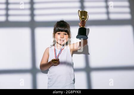 Dunkelhaariges Mädchen mit Sportmeisterschaftsmedaillen, das mit den Händen ihren Pokal über die Schatten eines Fensters mit einem Verschluss über ihr hebt. Sport und Stockfoto