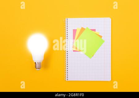 Ein Ideenkonzept - leuchtende Energiesparlampe und EIN Notizblatt mit Haftnotizen auf gelbem Tisch. Draufsicht, Flach Liegend, Leere Vorlage Stockfoto