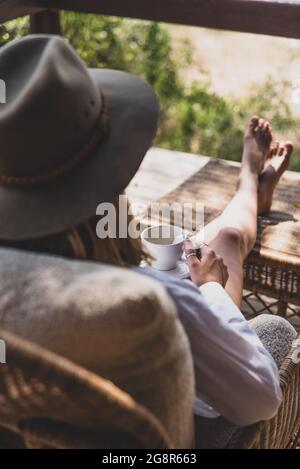 Eine Frau sitzt mit den Füßen nach oben und trinkt eine Tasse Tee, trägt einen Safarihut Stockfoto