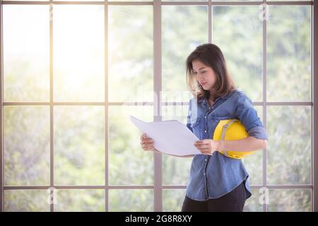 Asiatische schöne weibliche Ingenieurin in blauen Jeans-Shirt stehen und lesen Plan in Papier, halten gelben Schutzhelm mit Glasrahmen im Hintergrund Stockfoto