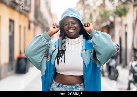 Coole junge afrikanische Frau mit Zöpfen, die lächeln und in der Stadt spazieren gehen. Sie trägt Freizeitkleidung und einen Hut. Stockfoto