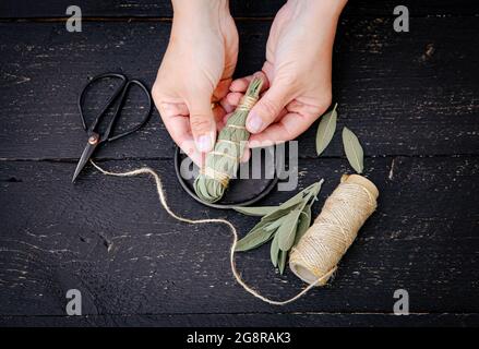 Person, die weißen Salbei Salvia apiana-Wischstock zu Hause mit hausgemachten Salbeiblättern macht. Baumwollstring und Vintage Schere zur Dekoration auf schwarz. Stockfoto