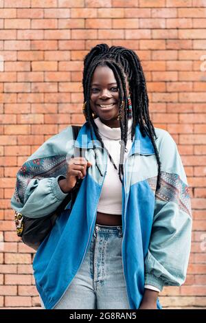 Glückliche und junge afrikanische Frau mit coolen Zöpfen, die lächeln und die Kamera betrachten. Sie trägt einen Rucksack. Stockfoto