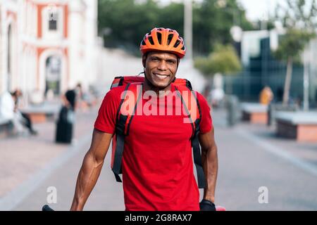 afroamerikanischer Lieferer, der Schutzhelm trägt, lächelt und schaut auf die Kamera auf der Straße. Stockfoto