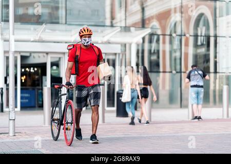 Afrikanischer Lieferer trägt Gesichtsmaske und trägt sein Fahrrad in der Stadt in der Art und Weise, wie er ein Paket liefert. Stockfoto