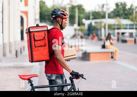Afrikanischer Lieferer mit Schutzhelm, der mit seinem Fahrrad in der Stadt steht und ein Paket hält. Stockfoto