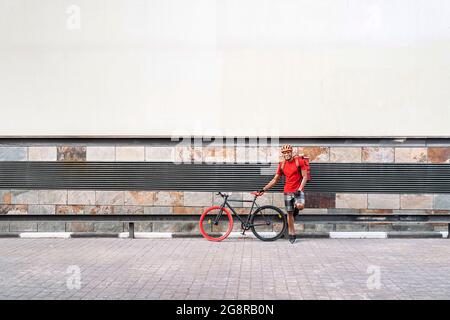 afroamerikanischer Lieferer mit Schutzhelm lächelt neben seinem Fahrrad und schaut auf die Kamera auf der Straße. Stockfoto