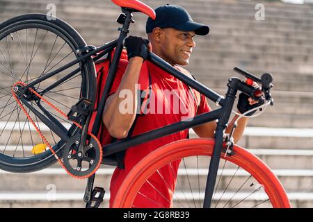Glücklicher Lieferer mit Mütze, der sein Fahrrad in der Stadt für die Lieferung eines Pakets im Weg hat. Stockfoto