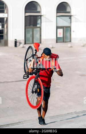 Afrikanischer Lieferer mit Mütze, der sein Fahrrad in der Stadt trägt, um ein Paket zu liefern. Stockfoto