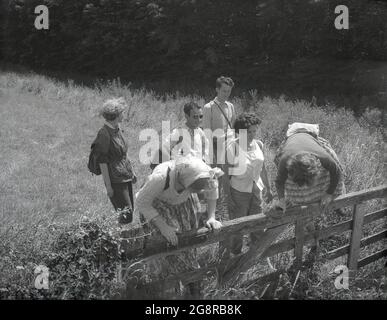 1950er Jahre, historisch, sommerlich und draußen auf dem Land auf den Noth-Downs in der Nähe von Nore, Kent, Engand, einige männliche und weibliche Wanderer stehen am Rande eines Feldes, an einem hölzernen Tor, wobei zwei der Damen versuchen, darüber zu klettern. Stockfoto