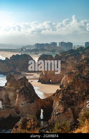 Wenn die Sonne untergeht, bilden natürliche Felsbögen und Klippen einen malerischen Blick auf den Strand, eine Stadt ist im Hintergrund sichtbar - Raum für Text Stockfoto