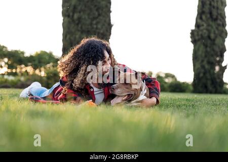 Junge afrikanerin mit lockigen Haaren, die im Park liegt und ihren reinrassigen Hund küsst. Stockfoto