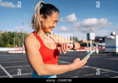 Glückliche aktive Frau mit Sportkleidung, die ihr Mobiltelefon verwendet und ihre Smart-Watch im Freien sucht. Stockfoto