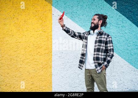 Glücklicher Hipster-Mann, der in der Stadt gegen eine bunte Mauer steht und mit seinem Handy ein Selfie macht. Stockfoto