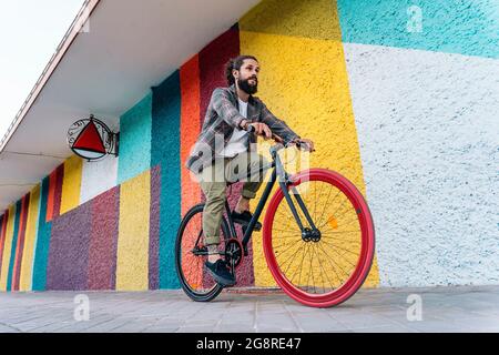 Junger Hipster-Mann mit großem Bart, der vor buntem Hintergrund auf dem Fahrrad reitet und nach vorne schaut. Stockfoto