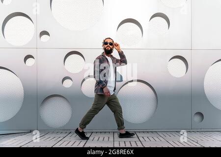 Junger Mann mit langem Bart und Sonnenbrille, der auf der Straße gegen die weiße Wand mit coolem Design läuft. Stockfoto
