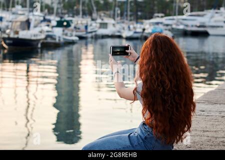 Junge Rotschopf-Frau macht mit ihrem Handy in einem Seehafen ein Foto Stockfoto