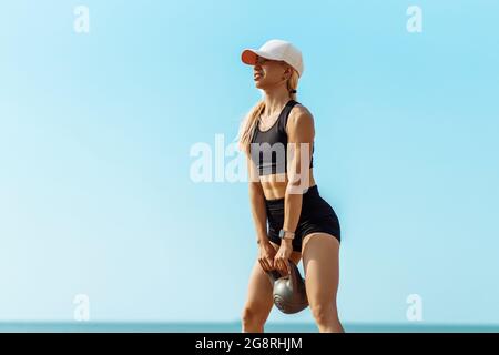 Sportlerin in Sportbekleidung mit Gewichten, sportliche Frau mit Kugelhantel beim Crossfit-Training am Strand am Morgen Stockfoto