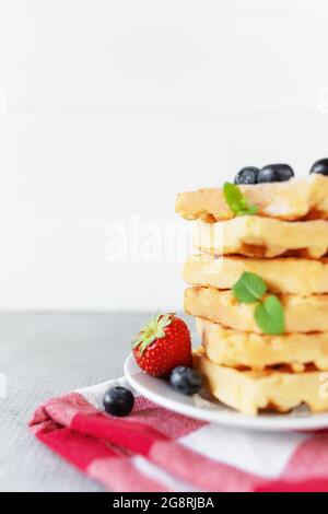 Weißer Teller auf rotem Küchentuch mit hausgemachten belgischen Waffeln, Heidelbeere, Erdbeerschnitt, Minzblatt, Zuckerpulver auf grauem Hintergrund Stockfoto