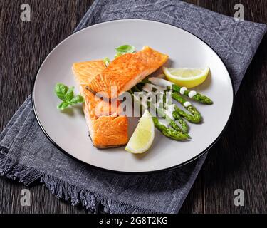 Lachsfilets mit Zitronenkeilen und gegrilltem Spargel, mit Sauce Hollandaise bestreut und auf einem Teller auf einem dunklen Holztisch Stockfoto