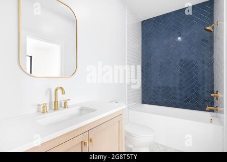 Ein wunderschön renoviertes Bad mit Schrank aus natürlichem Holz, goldenen Spiegeln und Wasserhähnen und einer blauen Duschwand mit Fischgrätmuster. Stockfoto