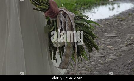 Nahaufnahme einer Hand der Bräute mit einem Blumenstrauß. Es gibt eine Pfütze mit Regenwasser auf dem Weg. Stockfoto