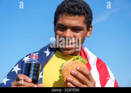 Afroamerikanischer Mann mit Flagge genießt Hamburger und Cola-Getränke im Freien Stockfoto